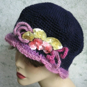 May include: A navy blue crocheted hat with a pink and white crocheted trim and a flower embellishment made of sequins.