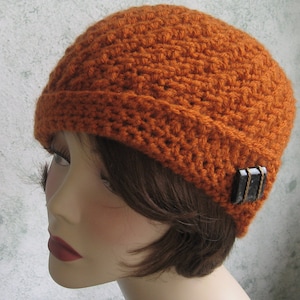 Può includere: Un cappello all'uncinetto di colore arancio bruciato con un bottone nero e argento sul lato.