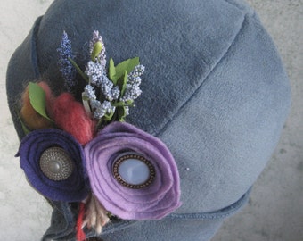May include: A blue felt cloche hat with a flower embellishment. The flower is made of purple, blue, and pink felt and has a small, round, blue button in the center.