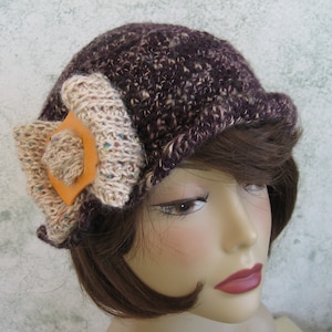 May include: A purple and brown knitted hat with a beige knitted flower accent. The flower has a brown leather center.
