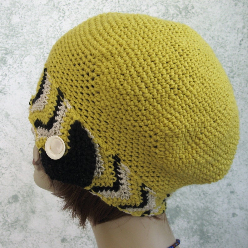 May include: A yellow crocheted hat with a black and white geometric pattern. The hat has a button on the side.