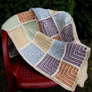 Puede incluir: Una manta de ganchillo con un diseño de patchwork que presenta cuadrados en varios colores, incluyendo morado, azul, verde, naranja y amarillo. La manta está hecha con hilo blanco y tiene un borde de ganchillo.
