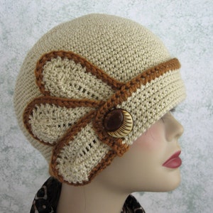 May include: A beige crocheted hat with a brown crocheted trim and a brown button. The hat has a unique design with a flower-shaped detail on the side.