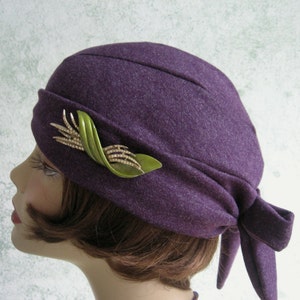 May include: A purple wool hat with a green and gold pin. The hat has a bow at the back.
