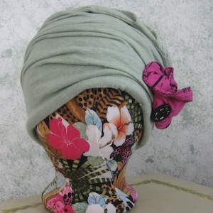 Op de afbeelding: Een turban-stijl hoed in saliegroen met een roze bloemenaccent van stof en een zwarte knoop.
