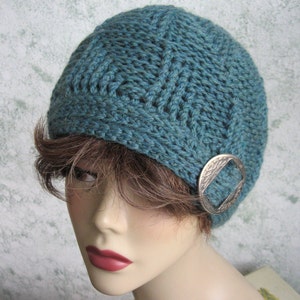 May include: Teal blue knitted hat with a silver metal buckle. The hat has a cable knit pattern.