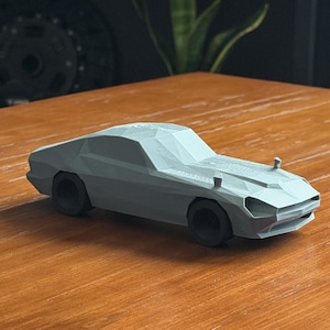 May include: A light gray, low-poly model of a classic sports car. The car has a sleek, angular design with black wheels and a detailed front end. The model is sitting on a wooden surface.