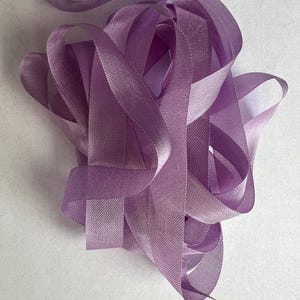 May include: A spool of lavender-colored ribbon with a satin finish. The ribbon is folded and twisted, showing its texture and sheen.