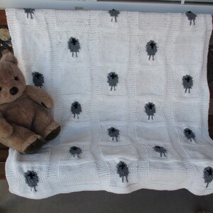 May include: White knitted baby blanket with a repeating pattern of gray sheep. The blanket is lying on a wooden surface with a brown teddy bear on top of it.