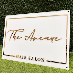 Puede incluir: Un letrero blanco con un borde dorado y el texto "The Avenue Hair Salon" en letras doradas.