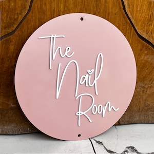 K&ouml;nnte beinhalten: Ein rundes Schild aus rosa Holz mit dem Text "The Nail Room" in wei&szlig;er Kursivschrift. Ein kleines wei&szlig;es Herz befindet sich &uuml;ber dem Wort "Nail".