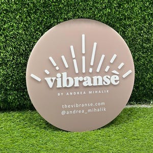 K&ouml;nnte beinhalten: Ein rundes, hellrosa Schild mit einem wei&szlig;en Sonnenstrahlen-Design und dem Text "vibranse" in Wei&szlig;, gefolgt von "BY ANDREA MIHALIK" in kleinerer wei&szlig;er Schrift. Unter dem Text stehen "thevibranse.com" und "@andrea_mihalik" in wei&szlig;er Schrift.