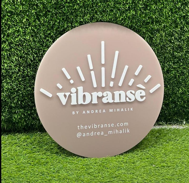 K&ouml;nnte beinhalten: Ein rundes, hellrosa Schild mit einem wei&szlig;en Sonnenstrahlen-Design und dem Text "vibranse" in Wei&szlig;, gefolgt von "BY ANDREA MIHALIK" in kleinerer wei&szlig;er Schrift. Unter dem Text stehen "thevibranse.com" und "@andrea_mihalik" in wei&szlig;er Schrift.