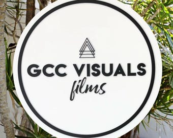 Puede incluir: Un letrero redondo blanco con borde negro. El letrero tiene un logotipo en blanco y negro con el texto "GCC VISUALS films" y un diseño de triángulo estilizado.