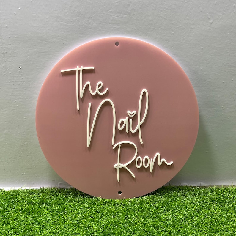 K&ouml;nnte beinhalten: Ein rundes rosa Schild mit dem Text "The Nail Room" in wei&szlig;er kursivschrift. Ein kleines wei&szlig;es Herz ist im Wort "Nail" enthalten.