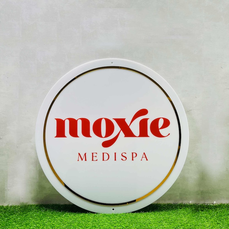 K&ouml;nnte beinhalten: Rundes wei&szlig;es Schild mit goldener Umrandung und dem Wort "moxie" in roten Buchstaben. Unter dem Wort "moxie" steht der Text "MEDISPA" in roten Buchstaben.