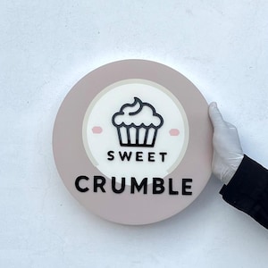 May include: A round, pink sign with a white circle in the centre. The circle has a black cupcake icon and the text "SWEET CRUMBLE".