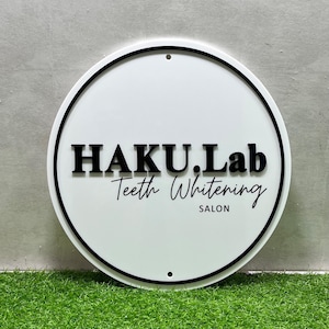 K&ouml;nnte beinhalten: Ein wei&szlig;es rundes Schild mit schwarzem Rand und dem Text "HAKU.Lab Teeth Whitening SALON" in Schwarz.