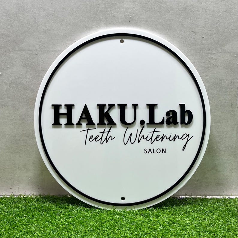 K&ouml;nnte beinhalten: Ein wei&szlig;es rundes Schild mit schwarzem Rand und dem Text "HAKU.Lab Teeth Whitening SALON" in Schwarz.