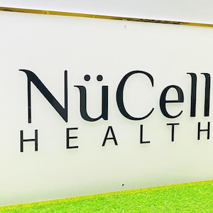 Op de afbeelding: Een rechthoekig bord met een gouden lijst en een witte achtergrond. Het bord bevat de woorden "NüCell HEALTH" in zwarte, gestileerde letters. Het bord wordt weergegeven op een bed van groen kunstgras.