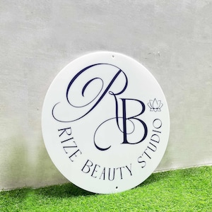 K&ouml;nnte beinhalten: Ein wei&szlig;es rundes Schild mit einem schwarz-wei&szlig;en Logo, auf dem "Rize Beauty Studio" steht, mit einem stilisierten "RB" in der Mitte und einem kleinen Lotusbl&uuml;ten-Design rechts neben dem "B".