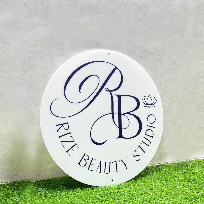 K&ouml;nnte beinhalten: Ein wei&szlig;es rundes Schild mit einem schwarz-wei&szlig;en Logo, auf dem "Rize Beauty Studio" steht, mit einem stilisierten "RB" in der Mitte und einem kleinen Lotusbl&uuml;ten-Design rechts neben dem "B".