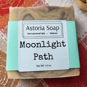 May include: A handcrafted vegan soap bar from Astoria Soap, labeled "Moonlight Path." The soap has a light brown and purple marbled design, wrapped with a light blue band. Net weight is 4.3 oz.