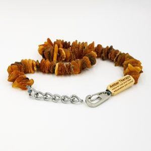 Pode incluir: Um colar de dentição de âmbar longo com uma corrente e um fecho prateados. O colar tem uma etiqueta de madeira com o texto "Amber Therapy" e uma breve mensagem sobre os benefícios do âmbar.