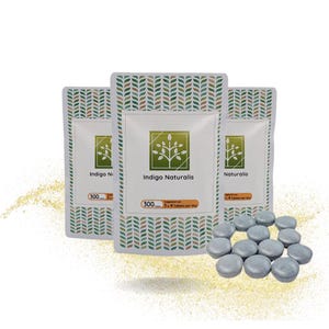 Op de afbeelding: Drie witte zakjes met een groen en bruin bladerpatroon, met het opschrift "Indigo Naturalis". De zakjes hebben een centraal vierkant met een boomlogo en de tekst "300". Een groep grijze tabletten ligt voor de zakjes.