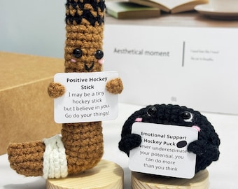 Puede incluir: Dos artículos de hockey de ganchillo: un palo de hockey marrón y un disco de hockey negro. El palo de hockey sostiene un letrero que dice "Positive Hockey Stick". El disco sostiene un letrero que dice "Emotional Support Hockey Puck".