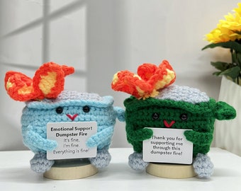Peut inclure: Deux jouets en peluche en crochet en forme de bennes à ordures avec des flammes sur le dessus. La benne bleue tient une pancarte qui dit "Emotional Support Dumpster Fire It's fine, I'm fine. Everything is fine!" La benne verte tient une pancarte qui dit "Thank you for supporting me through this dumpster fire!"