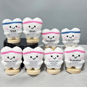 Pode incluir: Uma coleção de dentes de crochê feitos à mão com rostos sorridentes e várias mensagens. Cada dente tem um corpo branco, detalhes em rosa ou azul e uma base de madeira. As mensagens incluem "Pai", "Mãe" e "Filha".