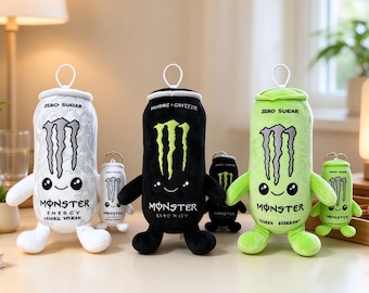 Puede incluir: Tres peluches con forma de latas de bebida energética en blanco, negro y verde claro, cada uno con cara sonriente, brazos, piernas y una anilla para colgar. Llaveros de peluche más pequeños a juego acompañan a cada peluche grande.