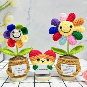 May include: Three crocheted flower pots with different messages. The first pot has a rainbow flower with a smiling face and the message "Thank you for helping me grow". The second pot has a rainbow heart with a smiling face and the message "Love is love". The third pot has a rainbow flower with a smiling face and the message "Emotional Support Plant Life can be tough sometimes, but remember so are you!"