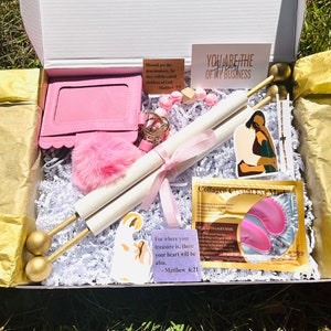 May include: A pink gift box filled with various items, including a pink wallet, a pink fluffy pom-pom, a white scroll with a pink ribbon, two gold-tipped sticks, a pink and white eye mask, and a small card with the text "For where your treasure is, there your heart will be also. - Matthew 6:21".