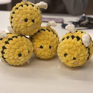 May include: Four crocheted bee plushies with black and yellow stripes, white wings, and black eyes. The plushies have pink cheeks and are stacked on top of each other.