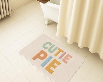 Puede incluir: Una alfombra de baño rectangular de color blanquecino con las palabras "CUTIE PIE" en letras de bloque coloridas. Las letras son en tonos naranja, azul, verde, rosa y amarillo. La alfombra está en un baño.