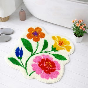 May include: A floral-shaped bath mat with a cream-colored background. The mat features colorful flowers in orange, yellow, pink, and blue, with green leaves and stems. The mat is placed on a white wooden floor, near a bathtub.