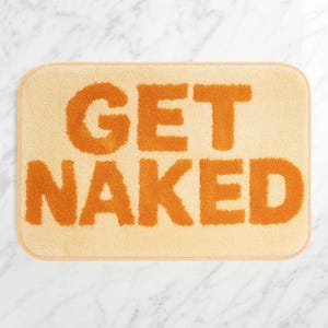 Può includere: Un tappetino da bagno rettangolare con angoli arrotondati. Il tappetino è di colore beige chiaro con le parole "GET NAKED" in grandi lettere arancioni e strutturate. Il tappetino ha un aspetto morbido e felpato.