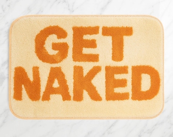 Op de afbeelding: Een rechthoekige badmat met afgeronde hoeken. De mat is lichtbeige van kleur met de woorden "GET NAKED" in grote, oranje, getextureerde letters. De mat heeft een zacht, pluche uiterlijk.