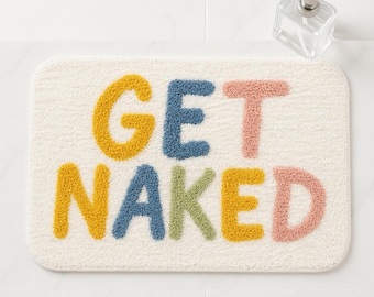 Puede incluir: Una alfombrilla de baño rectangular de color blanco roto con textura de mechones que presenta el texto GET NAKED en letras multicolores. Las letras son de color amarillo, azul, verde y rosa. Un dispensador de jabón de cristal transparente.