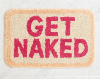 Op de afbeelding: Rechthoekige badmat met de woorden "GET NAKED" in felroze, gecentreerd op een beige achtergrond. De mat heeft een perzikkleurige rand en is gemaakt van een zacht, absorberend materiaal.