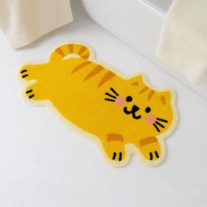 Op de afbeelding: Een gele katvormige badmat met oranje strepen en een lachend gezicht. De mat heeft een crèmekleurige rand en ligt op een witte tegelvloer. Het gezicht van de kat heeft roze wangen en zwarte snorharen.