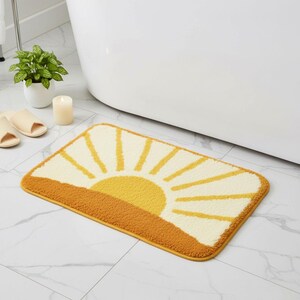 May include: A rectangular bath mat featuring a yellow sun design against a cream background. The sun's rays extend from a yellow-orange horizon. The mat has a yellow border and is placed on a white tile floor.