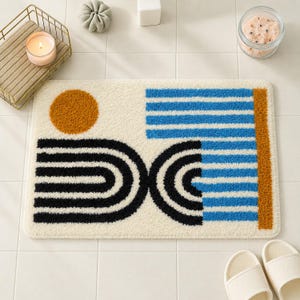 May include: A rectangular bath mat with a cream-colored base featuring a tufted design. The pattern includes a solid orange circle, black concentric semi-circles, and a section of horizontal blue stripes with a vertical orange border on the right.