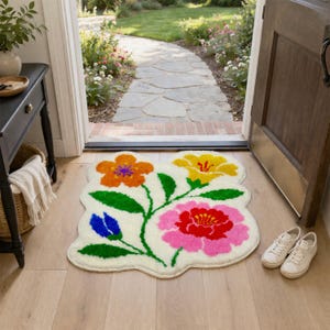 May include: A floral-shaped rug with a cream background and colorful flowers in orange, yellow, pink, and blue. The rug is placed inside a doorway, with white sneakers nearby. The rug's design features green stems and leaves.