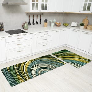 May include: Two kitchen rugs with an abstract pattern in green, yellow and blue. The rugs are positioned in front of white kitchen cabinets.