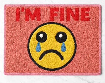 Puede incluir: Una alfombra rectangular con fondo rosa y borde rosa. El texto "I'M FINE" está en rojo en la parte superior. Un emoji amarillo con ojos negros, un ceño fruncido negro y lágrimas azules está en el centro.