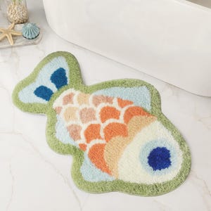 Può includere: Un tappetino da bagno a forma di pesce con una superficie testurizzata e trapuntata. Il design presenta un bordo verde, una coda con accenti blu scuro e un corpo decorato con squame sovrapposte nei toni dell'arancio, pesca e crema, con un occhio blu.