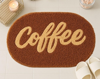 May include: Brown oval bath mat with the word "Coffee" in a light tan, cursive font. The mat has a textured, plush surface. White slippers are visible in the bottom right corner. A candle, soap, and other bathroom accessories are in the background.